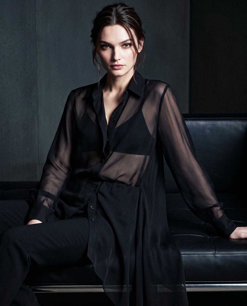 Model wearing a black sheer silk chiffon shirt and black tailored crepe trousers seated on a sofa in an editorial image by A|LOUD