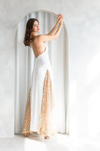 Back view of a model wearing an A|LOUD white floor-length gown with golden lace panels and low open back, posing beneath a minimalist arch.