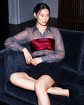 Model seated wearing the ÉLYR burgundy silk satin bandeau over a sheer charcoal silk chiffon shirt with a black mini skirt by A|LOUD