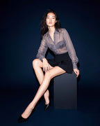 Model seated wearing the ÉLYR sheer charcoal silk chiffon shirt with black mini skirt in an editorial studio image by A|LOUD