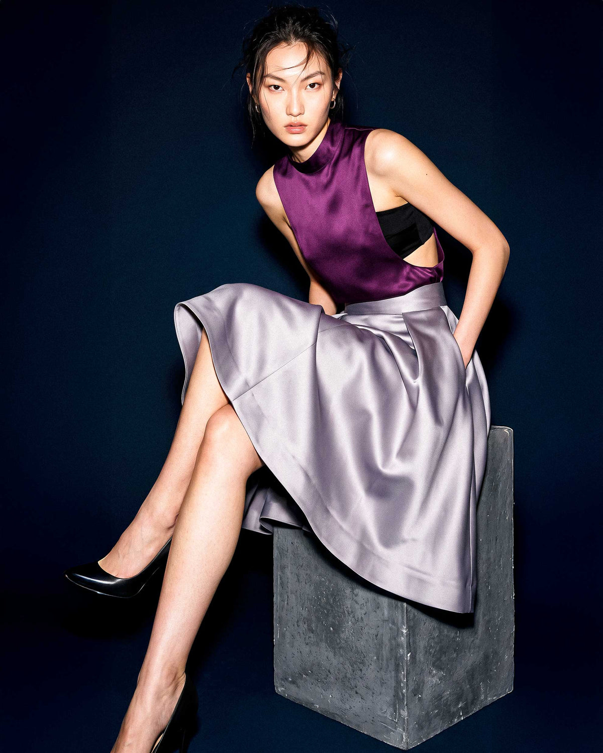 Model seated on a concrete plinth wearing the A|LOUD plum silk satin top and silver grey pleated midi skirt.