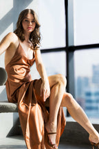 Model seated in an A|LOUD copper satin slip dress with thin straps, bathed in natural light with a city skyline in the background.
