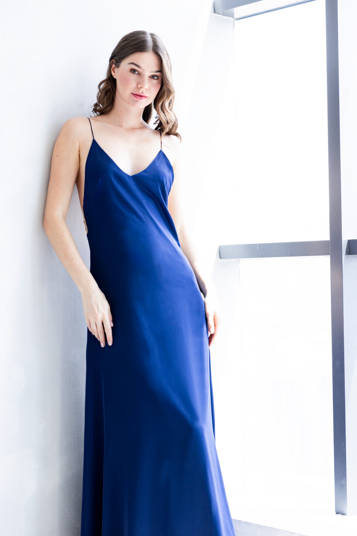 Model wearing an A|LOUD long blue satin gown with V neckline and thin straps, standing in soft natural light.
