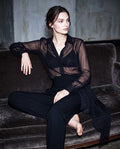 Model seated on a sofa wearing a black sheer silk chiffon shirt with black tailored crepe trousers by A|LOUD